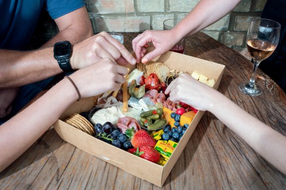 Hands reaching into a shared box of assorted fruits, cheeses, and snacks on a wooden table.