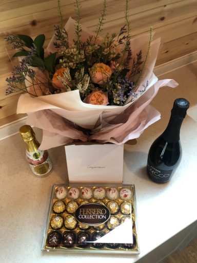 A gift arrangement featuring a floral bouquet, champagne bottle, and a box of chocolates.