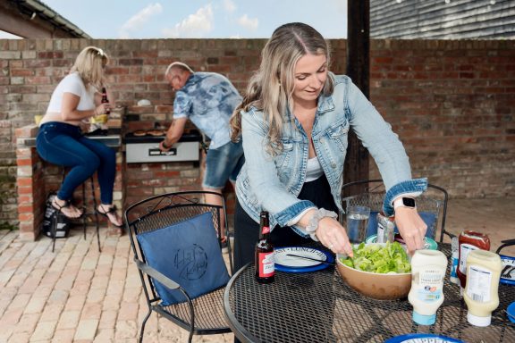 A woman prepares a salad at a barbecue gathering with two others in a garden setting.