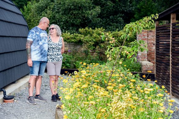 A couple stands together near vibrant yellow flowers in a garden setting.
